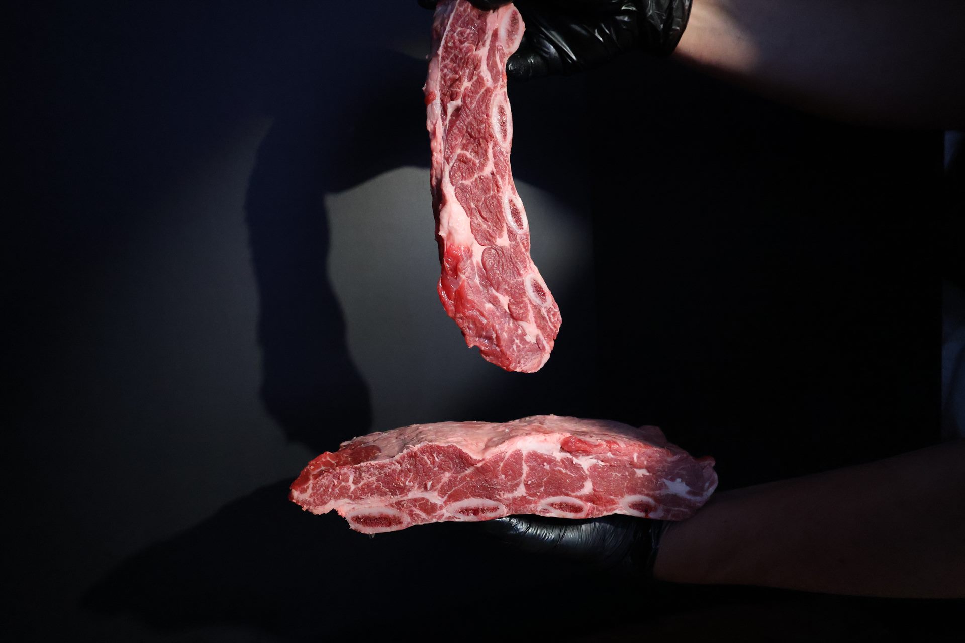 A butcher in black gloves holds up two bone-in beef short-rib strips against a dark backdrop