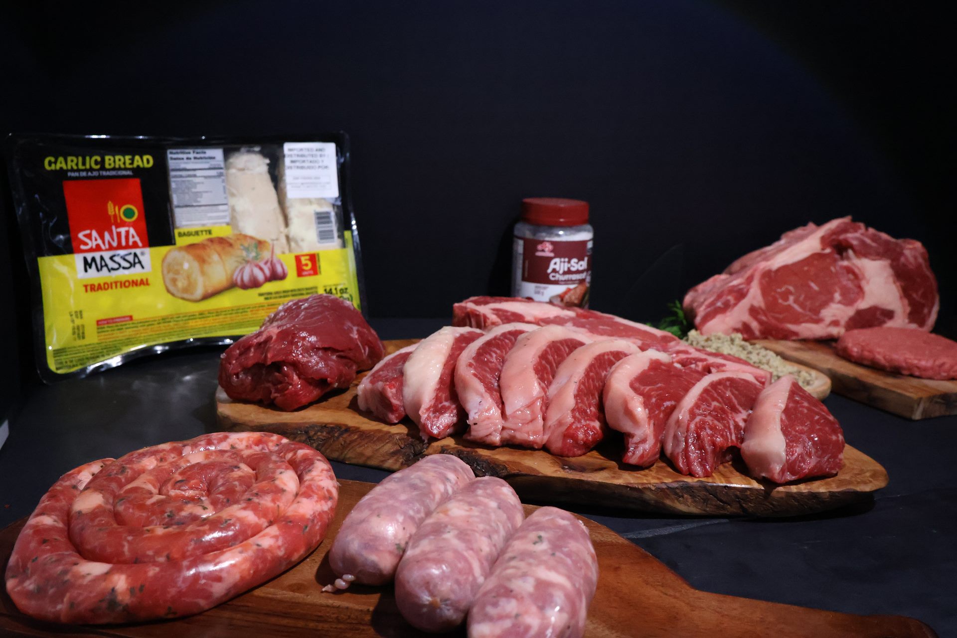 A churrasco spread on wooden boards: ribeye, sliced short rib, sausage coil and a garlic bread pack